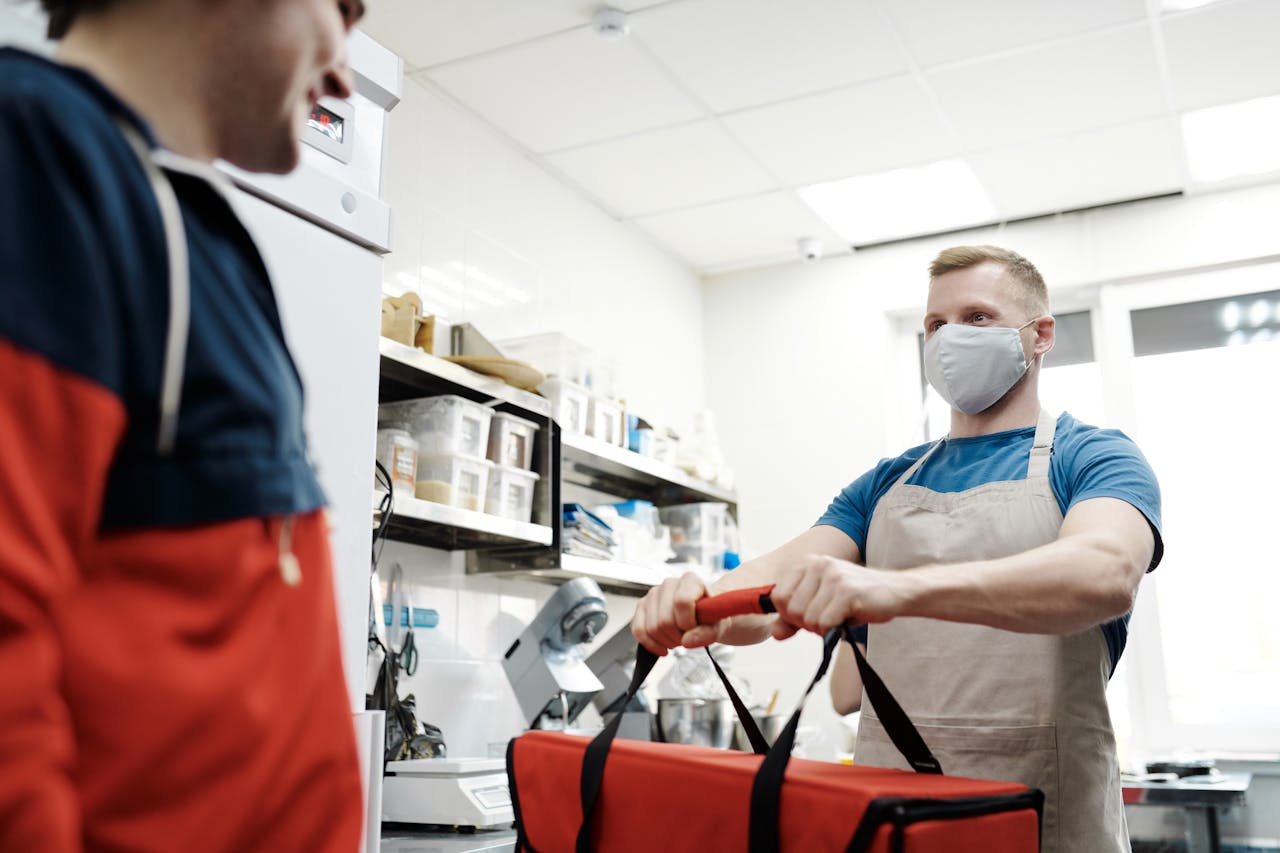 A male food delivery worker in an apron and face mask handling a thermal bag.